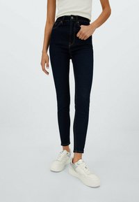 Tunna mörkblå jeans i denim med orange sömmar, hög midja och en slimmad passform, tillsammans med vita och grå sneakers.