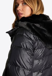 Schwarze Daunenjacke mit glänzender Oberfläche und einer Kapuze aus Kunstfell. Gestepptes Design mit sauber genähten Paneelen.