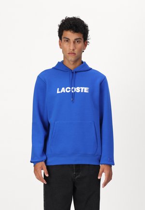 Sweat-shirt à capuche bleu en tissu doux, doté d'une poche frontale et d'un imprimé blanc "LACOSTE". Manches longues avec des côtes aux poignets.