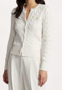 Mujer llevando un cárdigan blanco de punto trenzado con botones, y pantalones anchos blancos, con un pequeño logo bordado en el pecho.
