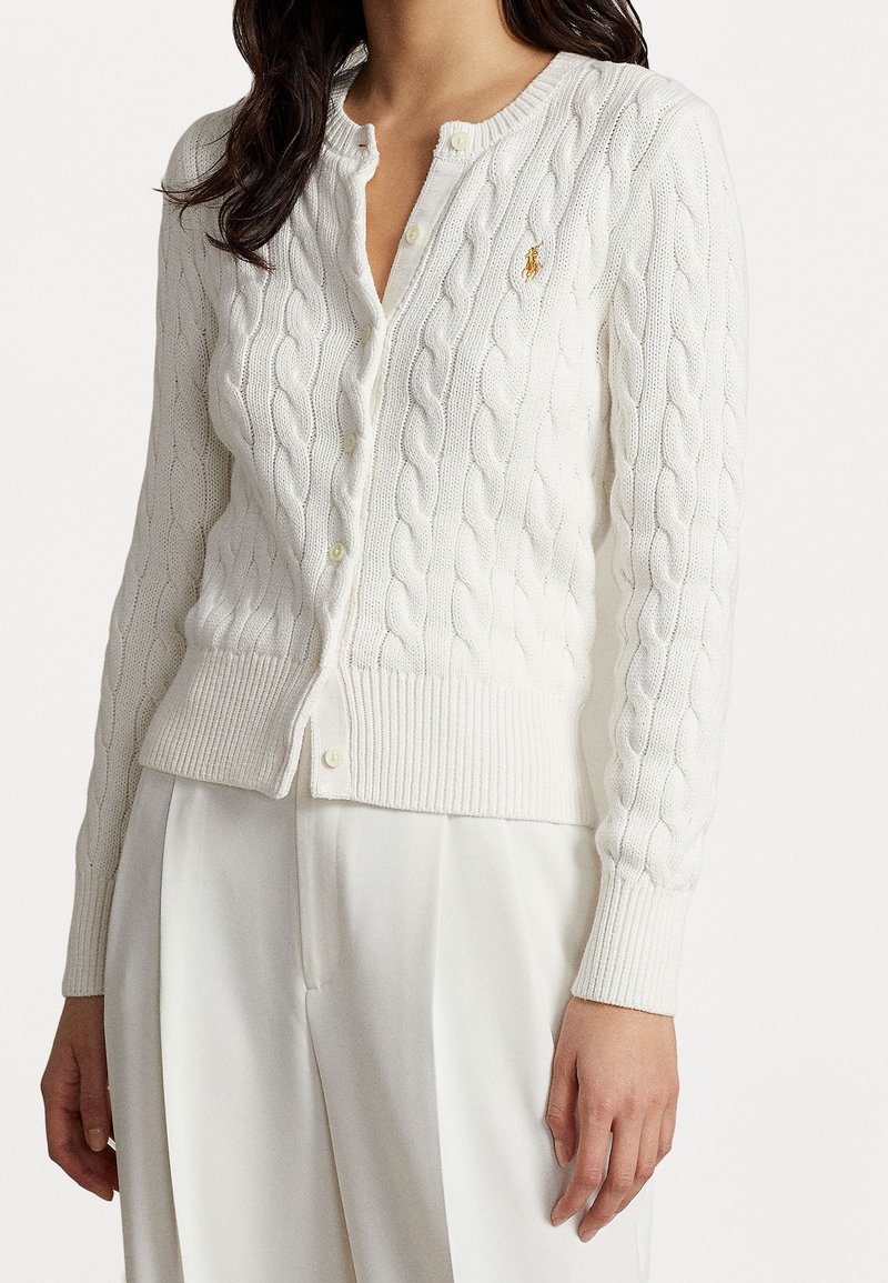 Mujer llevando un cárdigan blanco de punto trenzado con botones, y pantalones anchos blancos, con un pequeño logo bordado en el pecho.