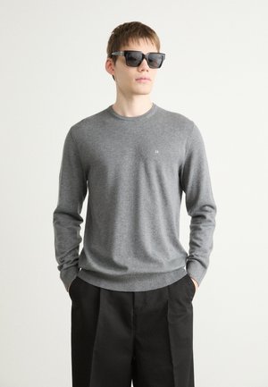 Gray knit sweater with long sleeves and a round neckline, featuring a small logo on the chest, paired with black trousers and sunglasses.