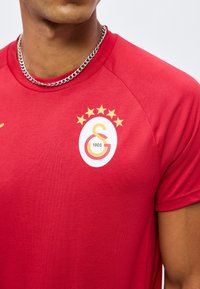 Άνδρας που φοράει κόκκινη αθλητική μπλούζα με το λογότυπο της Γαλατασαράι με πέντε αστέρια και ασημένιο κολιέ.