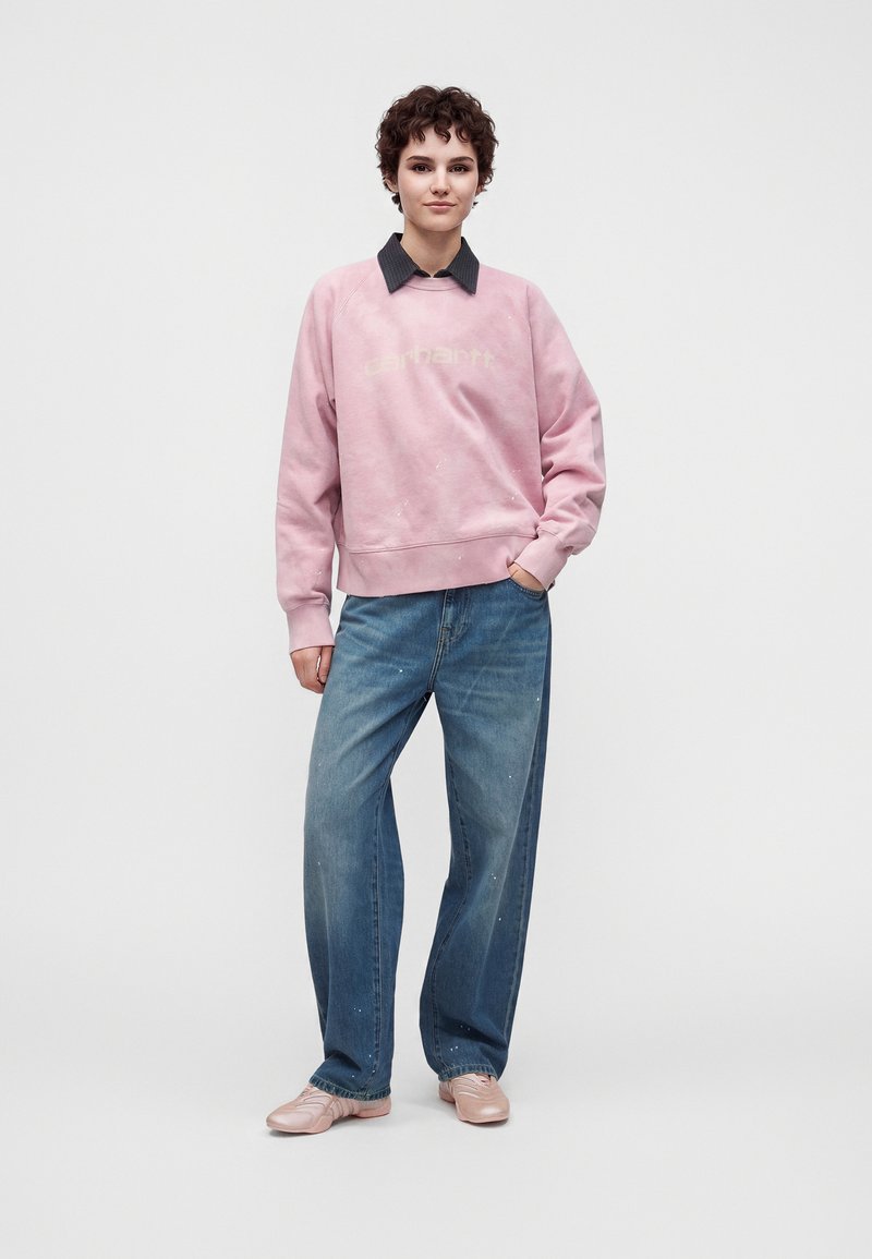 Junge Person mit kurzen lockigen Haaren, die einen pinken Pullover, weite blaue Jeans und pinke Schuhe trägt, steht mit einer Hand in der Tasche.