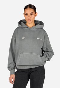 Sivá bavlnená mikina s predným vreckom, nastaviteľnou šnúrkou a rebrovanými manžetami. Obsahuje logo na hrudi a kontrastný text.