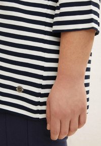 Navy- und weiß gestreiftes Oberteil aus geripptem Stoff. Verfügt über kurze Ärmel und ein silbernes Logo-Detail nahe dem Saum. Unten sichtbar ist navy.