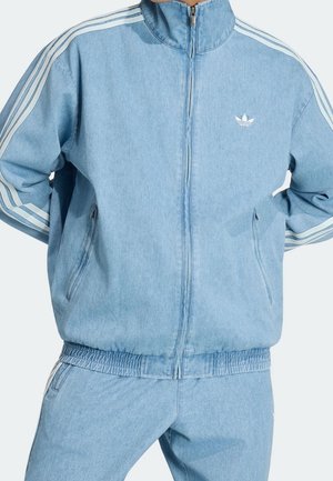 Personne portant un survêtement Adidas en denim bleu clair avec une veste zippée et trois bandes blanches le long des manches et des côtés du pantalon.