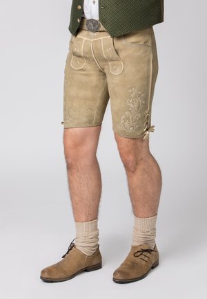 Beige Leder-Shorts in Kniehöhe mit gestickten floralen Mustern, frontalen Taschen und Schnürakzenten. Kombiniert mit beigen Strümpfen und braunen Schuhen.