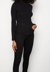 Mujer vestida con un suéter negro de cuello alto y manga larga, y jeans ajustados negros de cintura alta, con una mano en el bolsillo.