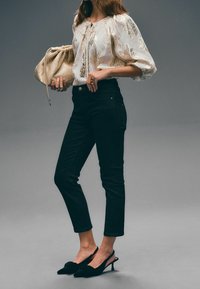 Bílé květinové blůzky s balónovými rukávy, černé úzké džíny a černé slingback podpatky. Modelka drží texturovanou béžovou tašku na stažení.