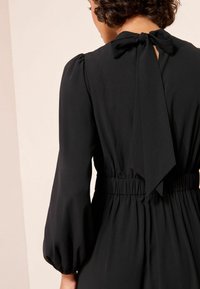 Melna kleita ar savilktu bantīti aizmugurē, puffīgu piedurkņu un elastīgu jostasvietu, kas nodrošina pieguļošu siluetu. Maigs, plūstošs audums.