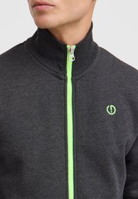 Chaqueta de cremallera de color gris oscuro con cuello alto, que presenta una cremallera verde y un acento de logo. La tela parece suave y tiene una textura lisa.