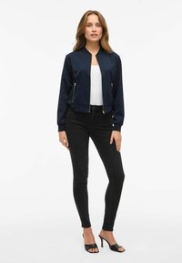 Veste de pilote navy avec fermeture éclair à l'avant et deux poches zippées dorées, associée à un jean noir ajusté et des talons ouverts noirs.