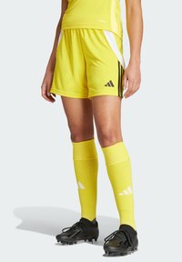 Gelbe Fußballshorts mit schwarzen Streifen, kombiniert mit gelben Kniestrümpfen. Schwarze Fußballschuhe mit Schnürsenkeln und einem aerodynamischen Design.