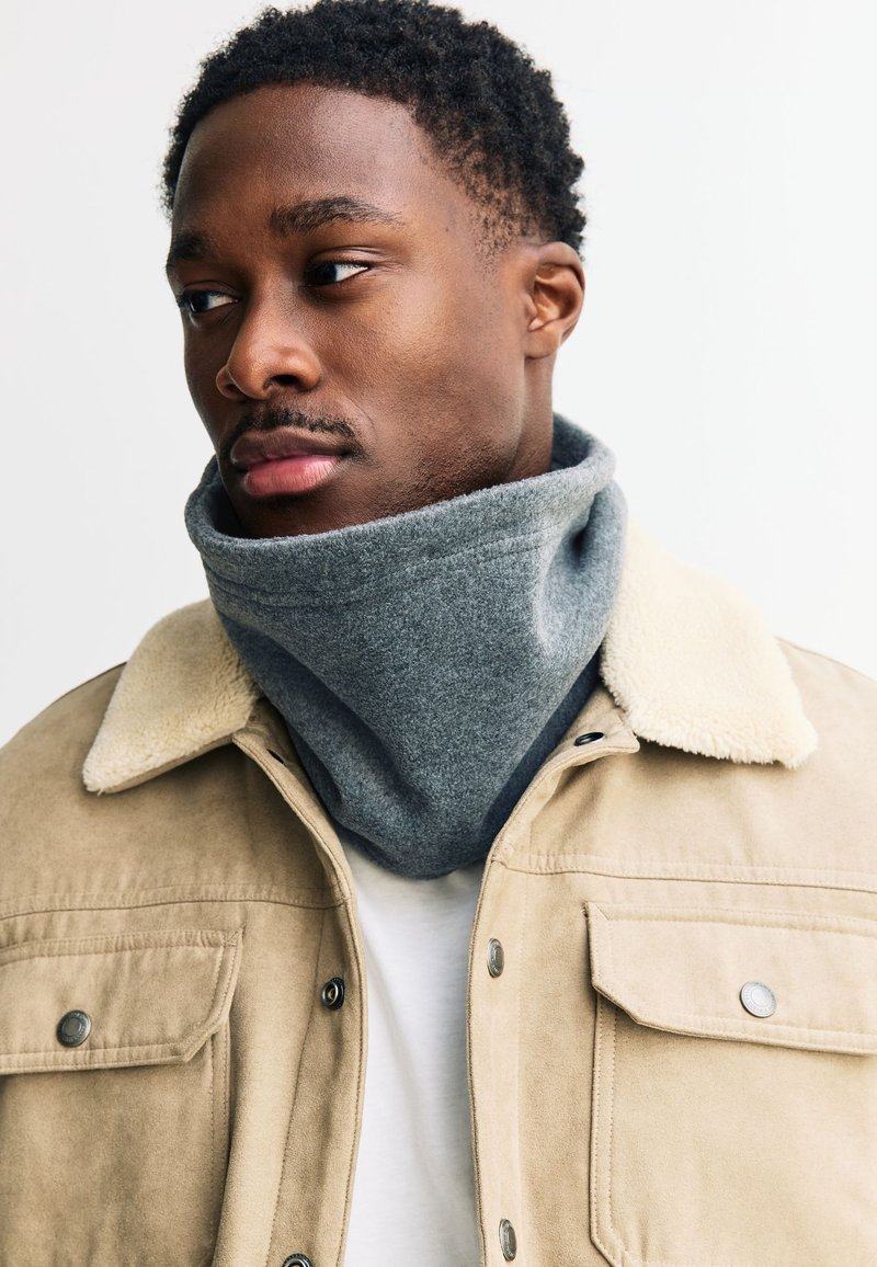 Szürke polár nyakmelegítő bézs dzseki alatt, amelynek szőrmés gallérja van, semleges háttér előtt. Egyszínű textúrájú és testre szabott kialakítású.