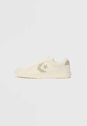 Lavt snøret cremefarvet sneakers med tekstureret krave, stjerne- og chevron-logo på siden, flad sål og snørebåndslukning på ensfarvet baggrund.