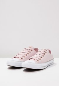 Paire de baskets basses en toile rose clair avec semelles en caoutchouc blanches et œillets en or rose, posées sur une surface blanche.