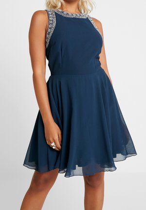 Frau trägt ein ärmelloses, marineblaues Kleid mit verzierter Halslinie und tailliertem Schnitt, ausgestelltem Rock, steht mit einer Hand, die das Kleid hält.