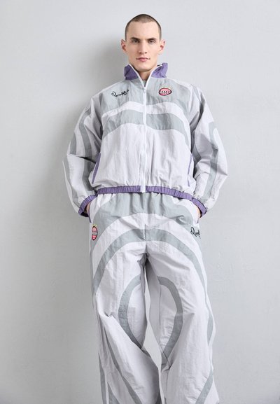 Light gray tracksuit featuring curved gray stripes, purple accents, and a round logo patch. Made from a lightweight material with elastic cuffs.