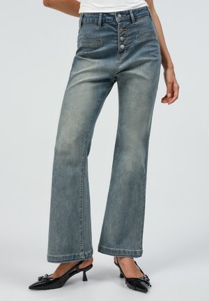 Femme portant un jean bleu délavé taille haute avec une braguette à boutons et des chaussures slingback noires à talons bas, debout avec une main détendue.