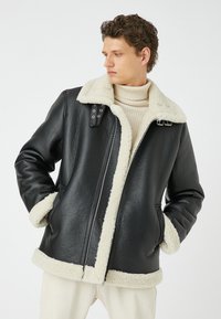 Giubbotto di pelle nera con colletto e polsini in shearling crema, caratterizzato da una chiusura zip frontale e dettagli con cinturino sulle spalle.