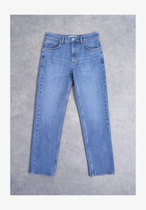 Jean en denim bleu avec jambes droites, fermeture avant à boutons et design à cinq poches, posé à plat sur une surface en béton gris.