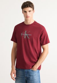 Bordo pamučna majica s okruglim izrezom, kratkim rukavima i velikim sivim logotipom "Calvin Klein" na prednjoj strani. Mekan materijal.