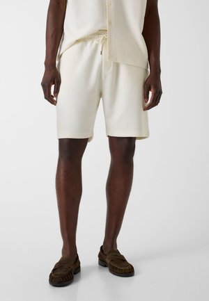 Man draagt crèmekleurige shorts met trekkoord en een bijpassend shirt met bruine loafers, staand tegen een effen lichte achtergrond.