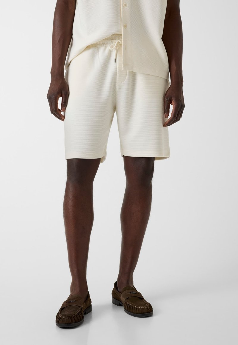 Homme portant un short crème avec cordon de serrage et une chemise assortie avec des mocassins marron, debout devant un fond clair uni.