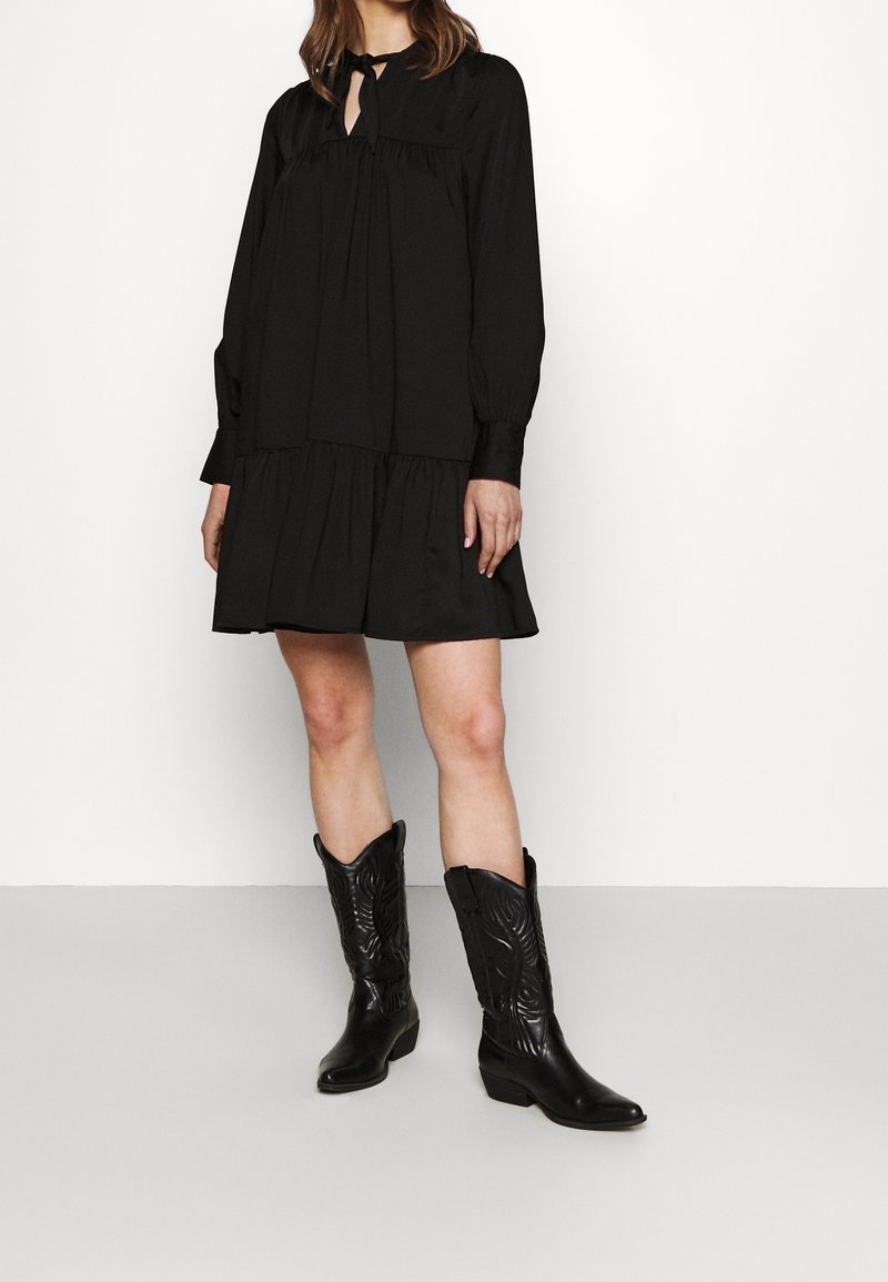 Vestido negro de capas con mangas largas y detalle de lazo en el cuello, combinado con botas de vaquero de cuero negro con costuras decorativas.