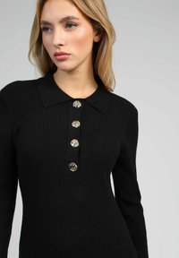 Robe noire en tricot côtelé avec un col, dotée d'une patte de boutonnage en accents métalliques. Silhouette ajustée et manches longues.