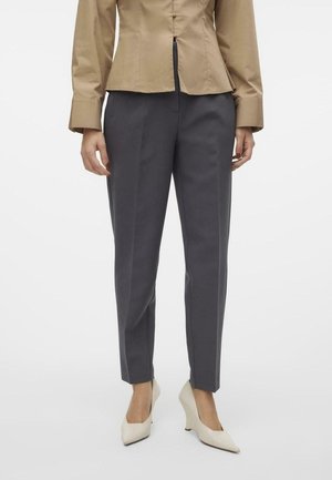 Femme portant un pantalon gris sur mesure, une chemise beige avec de longues manchettes, et des escarpins blancs à talons compensés bas et bouts pointus, debout devant un fond blanc.