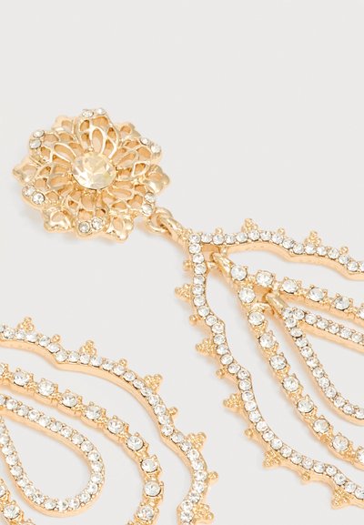 Aranyszínű filigrán fülbevalók áttetsző strasszkövekkel, virág és csepp alakban, világos háttéren.