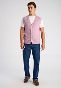 Hellrosa gestrickte Weste mit Zopfmuster, mit V-Ausschnitt und fünf Knöpfen, getragen über einem weißen T-Shirt und kombiniert mit blauen Jeans.