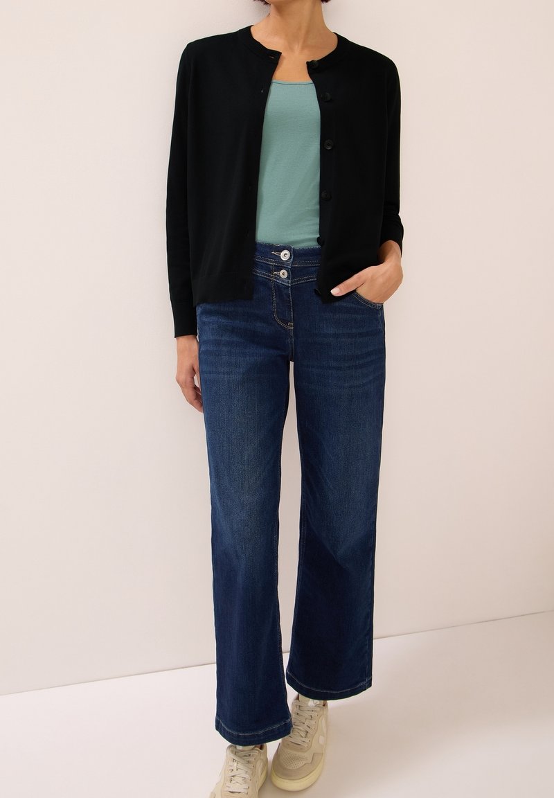 Femme portant un cardigan noir boutonné, un haut vert clair, un jean large bleu foncé et des baskets beige, debout avec une main dans la poche.