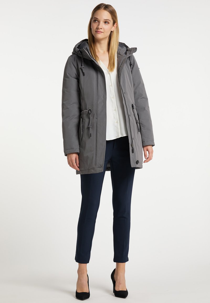 Graue gepolsterte Jacke mit Kapuze, ausgestattet mit Kordelzügen in der Taille, einem Reißverschluss und seitlichen Taschen. Kombiniert mit marineblauen Hosen und schwarzen Absätzen.