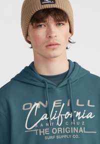 Tyrkysová mikina s kapucňou a sťahovacou šnúrkou, s grafickým potlačou, ktorá obsahuje nápis "O'Neill California" v bielej farbe. Nosená s pieskovou rebrovou čiapkou.