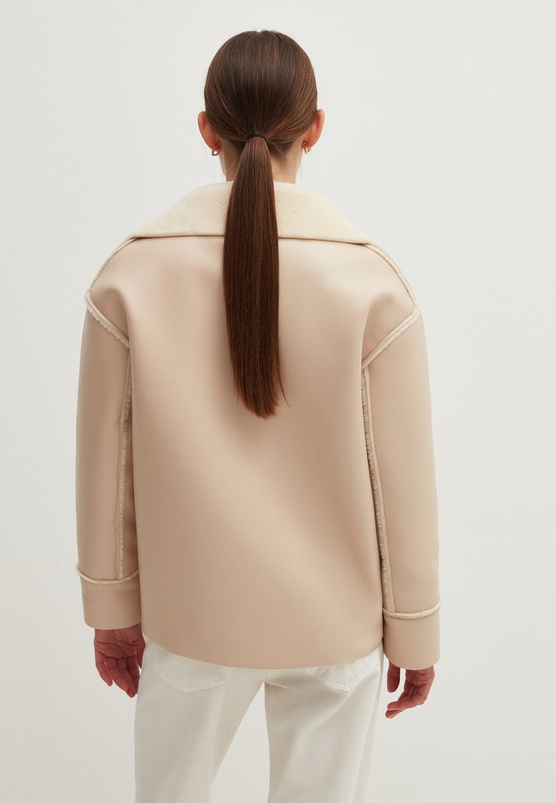 Beige Kunstlederjacke mit breitem Kragen, gefüttertem Innenfutter und sichtbaren Nähten. Über weißen Hosen getragen, die eine lockere Passform zeigen.