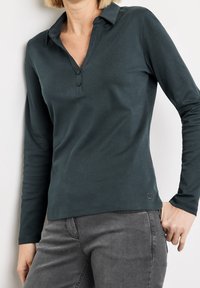 Mörkgrön långärmad pikétröja med krage, utrustad med en tre-knapps placket och en subtil logotypdetalj vid fållen. Mjuk tygtextur.
