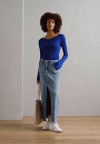 Langermet blå topp kombinert med et høyt splittet denimskjørt. Modellen bærer en beige tote og har på seg hvite sneakers, stående på tregulv.