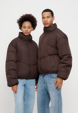 Deux jeunes adultes portant des doudounes marron foncé et des jeans bleu clair se tiennent côte à côte devant un fond uni.