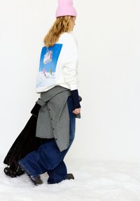 White sweatshirt with a blue graphic of skiers on the back, paired with loose denim pants and a black and white checkered shirt. Pink beanie on head.