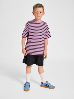 Junger Junge steht und trägt ein violett-schwarz gestreiftes T-Shirt, schwarze Shorts, weiße Socken und blaue Turnschuhe vor einfarbigem Hintergrund.