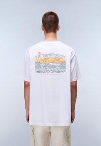 Vit bomullst-shirt med en grafisk design av berg och träd i grått och orange på ryggen, med korta ärmar och rund halsringning.