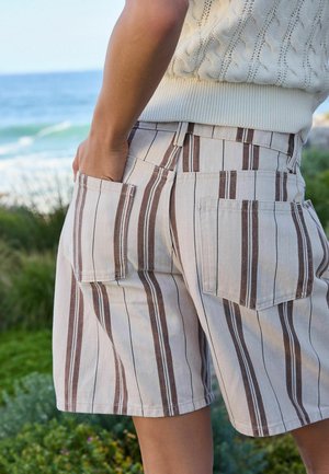 Person iført beige og brune vertikale stribede shorts med baglommer, stående tæt på grønt område og en krop af vand i baggrunden.