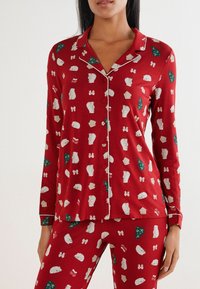 Červený set pyžama obsahující košili na knoflíky a odpovídající kalhoty, zdobený bílými medvědy, zelenými stromy a mašlemi v hravém vzoru.