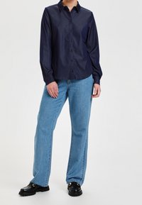 Camisa de botones azul marino de manga larga, con un cuello clásico. Combinada con vaqueros de pierna ancha en azul claro y mocasines negros.
