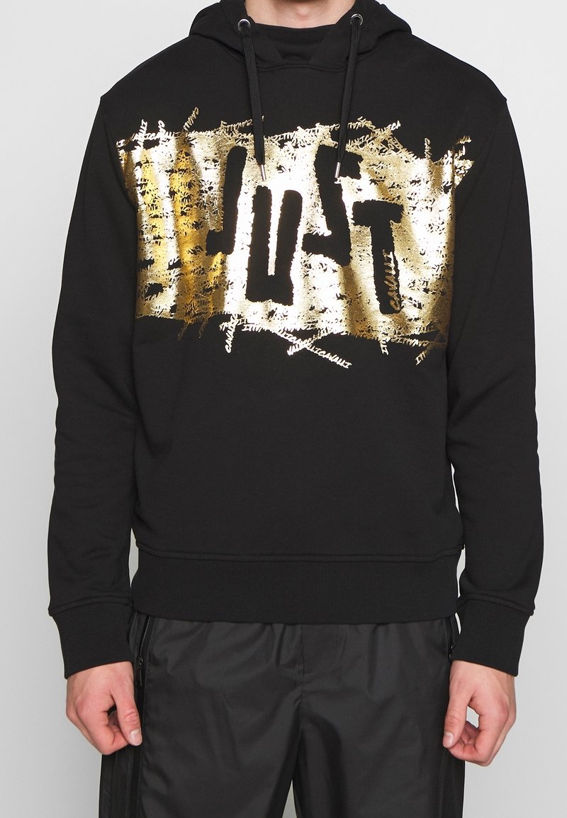 Sudadera con capucha negra hecha de algodón, con un gran estampado metálico dorado que dice "JUST" en la parte delantera. Incluye cordones ajustables y un dobladillo acanalado.