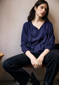 Blusa blu navy con scollo a V, spalle arricciate e maniche lunghe. Abbinata a jeans neri aderenti e scarpe nere. Tessuto liscio.