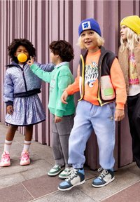 Quattro bambini si trovano di fronte a un muro texturizzato viola, indossando abbigliamento colorato. Sono evidenti una giacca con paillettes, felpe e sneakers distintive.